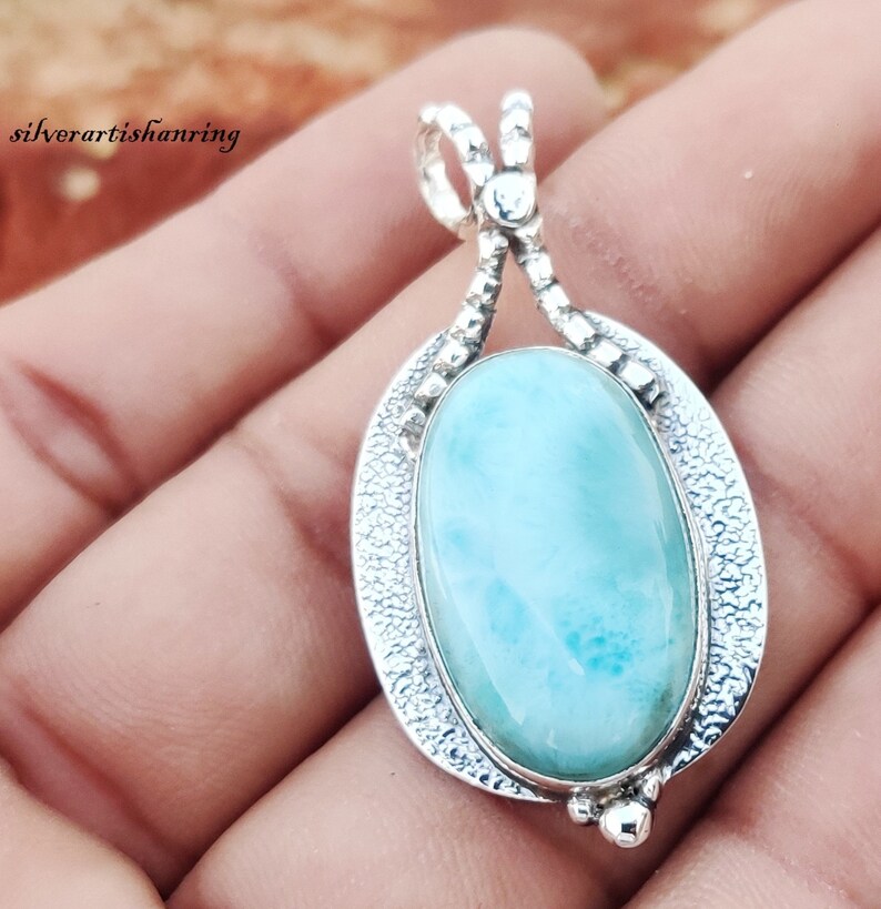 May include: A silver pendant with a light blue oval-shaped gemstone. The pendant has a detailed silver design around the gemstone.