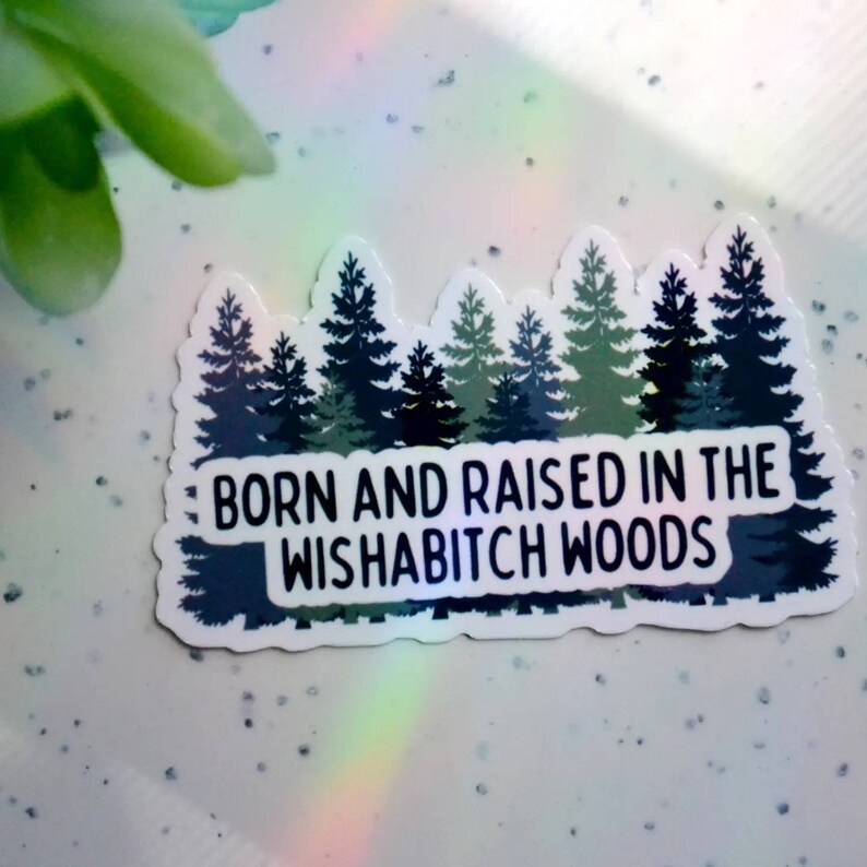 May include: A white sticker with a black outline and a forest silhouette. The text "Born and raised in the Wishabitch Woods" is printed in black.