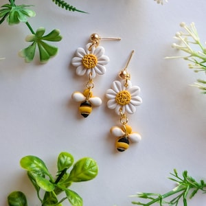 May include: A pair of dangle earrings featuring white daisy flowers with yellow centers and small, detailed bumblebees. The earrings have gold-colored posts and connecting chains. The bees have black and yellow stripes.