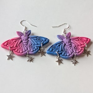 May include: A pair of handmade moth earrings. Each earring features a gradient of pink and blue wings with silver star charms. The earrings are made of polymer clay and have silver-colored ear wires.