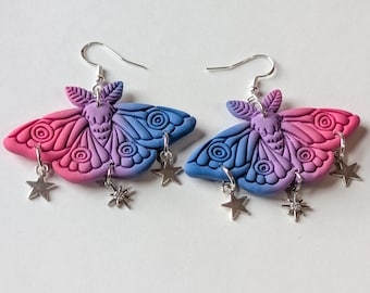 May include: A pair of handmade moth earrings. Each earring features a gradient of pink and blue wings with silver star charms. The earrings are made of polymer clay and have silver-colored ear wires.