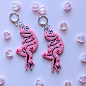 May include: A pair of pink frog earrings with red heart and white dot details. The earrings have silver hoops and are set against a light pink background with heart-shaped beads.