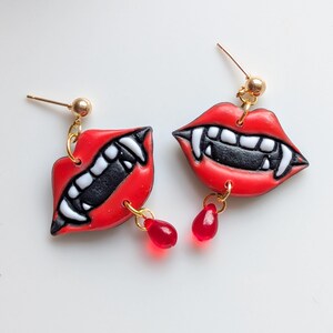 May include: A pair of novelty earrings featuring red lips with white fangs and black interiors. Each earring has a small, red, teardrop-shaped charm dangling below. The earrings have gold-colored posts and connecting rings.