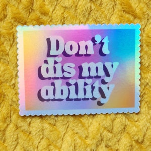May include: A holographic sticker with a scalloped edge. The sticker features the text "Don't dis my ability" in a white, retro font with heart accents. The background has a gradient of pink, purple, and blue hues.