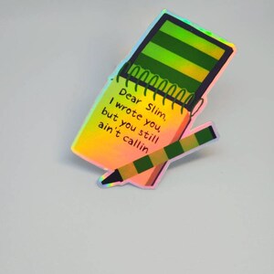 Puede incluir: Pegatina hologr&aacute;fica con un dise&ntilde;o de bloc de notas y l&aacute;piz. El bloc de notas tiene rayas verdes y amarillas y el texto "Dear Slim, I wrote you, but you still ain't callin". El l&aacute;piz es verde y marr&oacute;n.