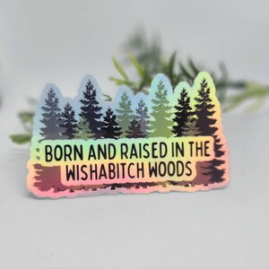 May include: A holographic sticker with a rainbow gradient background featuring a silhouette of a forest with the text "Born and raised in the Wishabitch Woods".