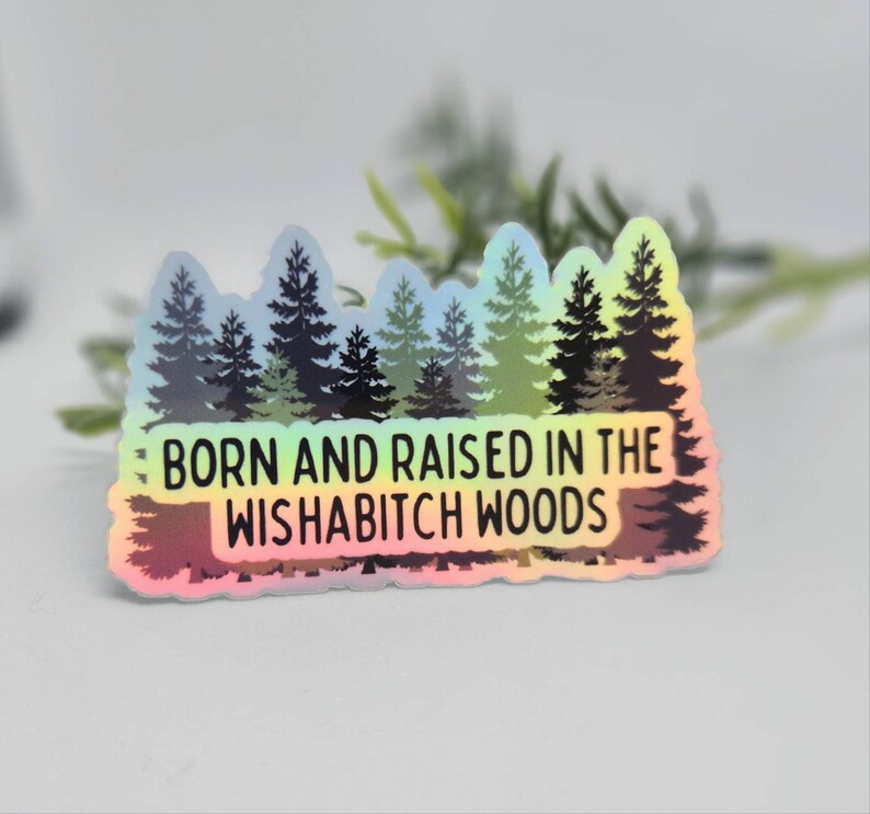 May include: A holographic sticker with a rainbow gradient background featuring a silhouette of a forest with the text "Born and raised in the Wishabitch Woods".