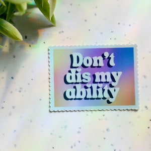 May include: A rectangular sticker with a scalloped edge features the text "Don't dis my ability" in white, bold font. The background has a gradient of pink, purple, and blue hues. The sticker is on a white surface.