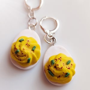 May include: A pair of silver earrings shaped like deviled eggs. The earrings are made of polymer clay and feature a yellow yolk with green chives and red paprika.