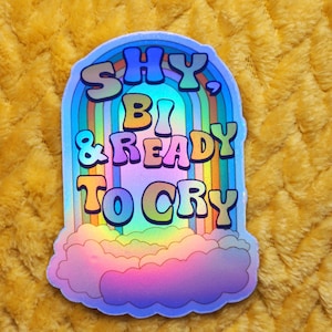 May include: A holographic sticker with a rainbow design and the text "SHY, BI & READY TO CRY" in a retro font.