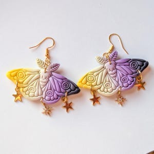 May include: A pair of handmade moth earrings. Each earring features a gradient of yellow, white, purple, and black, with gold-colored star and crystal charms dangling from the bottom. The earrings have gold-colored ear wires.