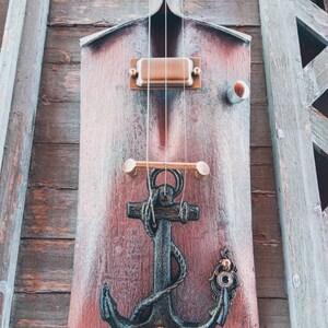 May include: A wooden shovel with a black anchor and a copper pickup attached to it. The shovel is painted red and has a distressed look. The anchor is a decorative element and the pickup is a musical instrument component.