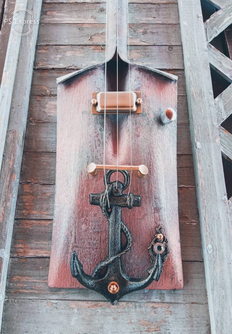 May include: A wooden shovel with a black anchor and a copper pickup attached to it. The shovel is painted red and has a distressed look. The anchor is a decorative element and the pickup is a musical instrument component.