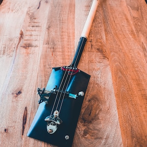 May include: A black shovel guitar with a wooden handle. The guitar has a metal tuning peg and a metal bottle opener.