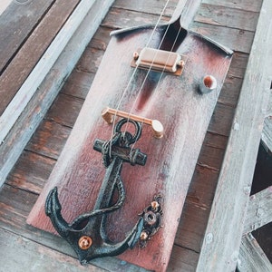 May include: A wooden guitar with an anchor design and a copper-colored finish. The guitar has a single pickup and a bridge with a metal plate. The anchor is black with gold accents.