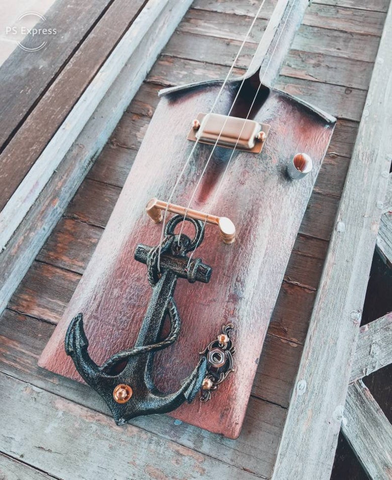 May include: A wooden guitar with an anchor design and a copper-colored finish. The guitar has a single pickup and a bridge with a metal plate. The anchor is black with gold accents.