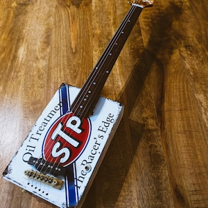 May include: A unique, handmade guitar with a body crafted from a vintage STP Oil Treatment sign. The guitar features a wooden neck, a skull design on the headstock, and the text "The Racer's Edge". The instrument rests on a wooden surface.
