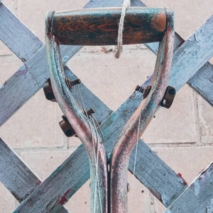 May include: A close-up of the top of a vintage shovel handle, painted in shades of blue, green, and copper. The handle is made of wood and has a rope tied around it. The shovel is hanging from a wooden lattice fence.