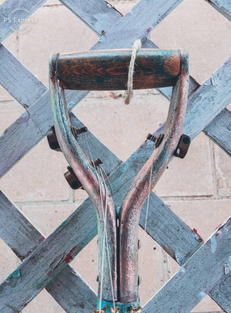 May include: A close-up of the top of a vintage shovel handle, painted in shades of blue, green, and copper. The handle is made of wood and has a rope tied around it. The shovel is hanging from a wooden lattice fence.