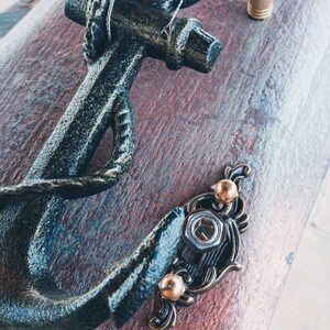 May include: A black metal anchor with a rope detail, mounted on a wooden board with a distressed finish. The anchor is attached with decorative metal accents and screws.