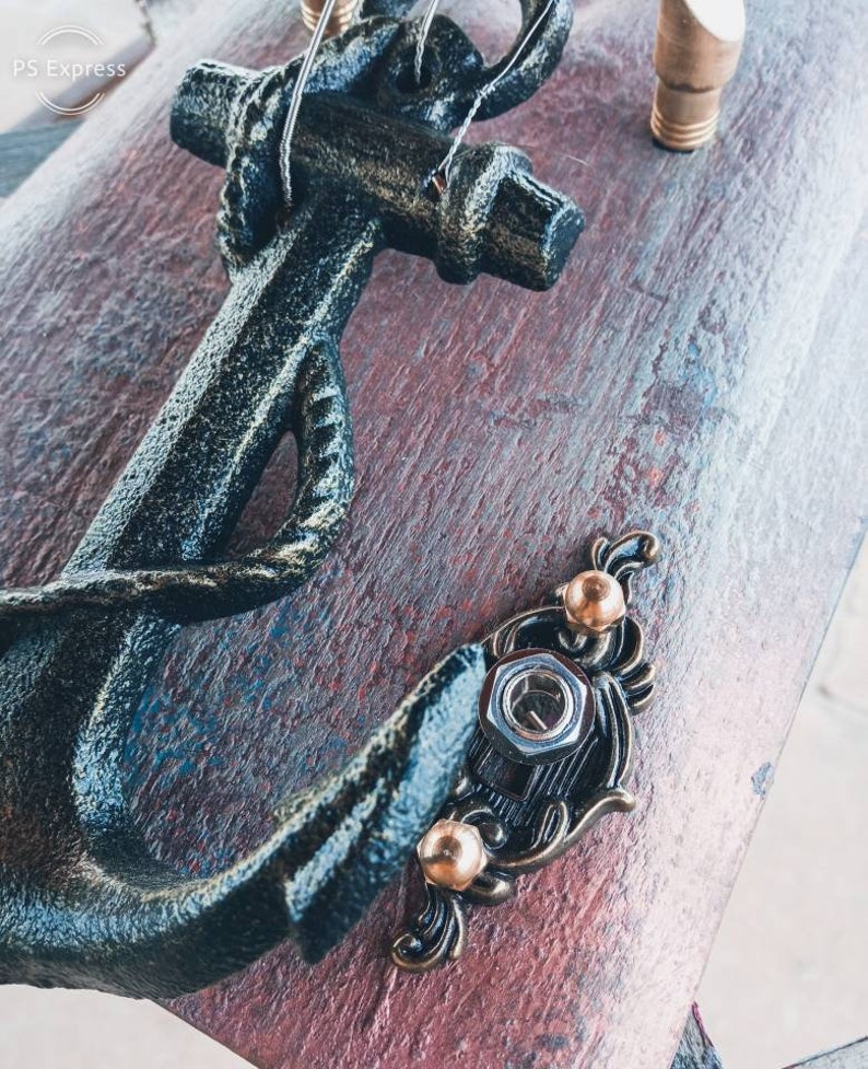 May include: A black metal anchor with a rope detail, mounted on a wooden board with a distressed finish. The anchor is attached with decorative metal accents and screws.