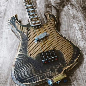 May include: A custom-made bass guitar with a unique, distressed wood finish. The body features a perforated design with a dark, weathered appearance. The neck has frets and strings, and the instrument has tuning knobs and pickups.