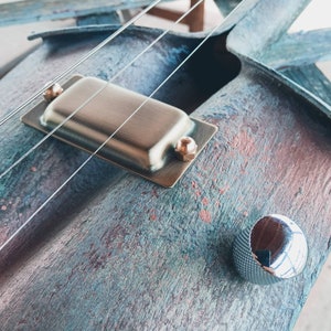 May include: Close-up of a custom-made electric guitar with a unique, rustic finish. The guitar features a single pickup, a silver knob, and two strings. The body of the guitar is made of wood with a blue, green, and brown color scheme.
