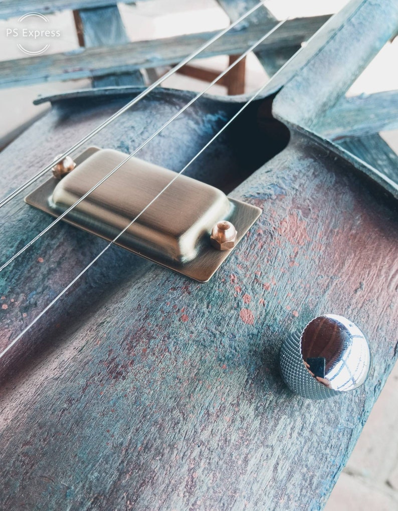 May include: Close-up of a custom-made electric guitar with a unique, rustic finish. The guitar features a single pickup, a silver knob, and two strings. The body of the guitar is made of wood with a blue, green, and brown color scheme.