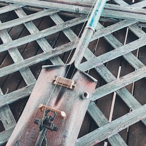 May include: A repurposed shovel turned into a unique musical instrument with an anchor design and a light blue neck. The instrument is resting on a wooden lattice.