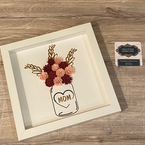 Könnte beinhalten: Ein gerahmtes Papierhandwerk mit einem Einmachglas, das mit rosa und burgunderfarbenen Papierblumen gefüllt ist. Das Glas hat ein Herz mit dem Wort "MOM" darin. Der Rahmen ist weiß und hat einen goldenen Rand.