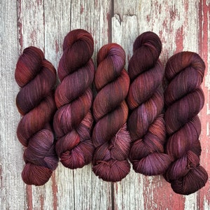 Puede incluir: Cinco ovillos de hilo en tonos de rojo oscuro, burdeos y morado. El hilo está retorcido y tiene un patrón de color variado.