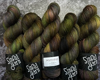 Könnte beinhalten: Fünf Knäuel grüner, brauner und lilafarbener Wolle mit dem Etikett "Sinful Yarn" und "Mystified" auf den Knäueln. Die Wolle ist ein Fingering-Gewicht und besteht aus 75% supergewaschener Merinowolle und 25% Nylon.