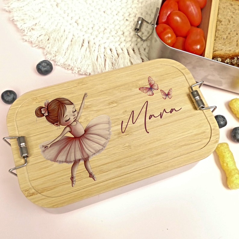 Pu&ograve; includere: Un portapranzo in legno con un'illustrazione di ballerina e il nome "Mana" in corsivo. La ballerina indossa un tut&ugrave; rosa e sta ballando. Due farfalle sono anche sul coperchio. Il portapranzo ha chiusure in metallo.