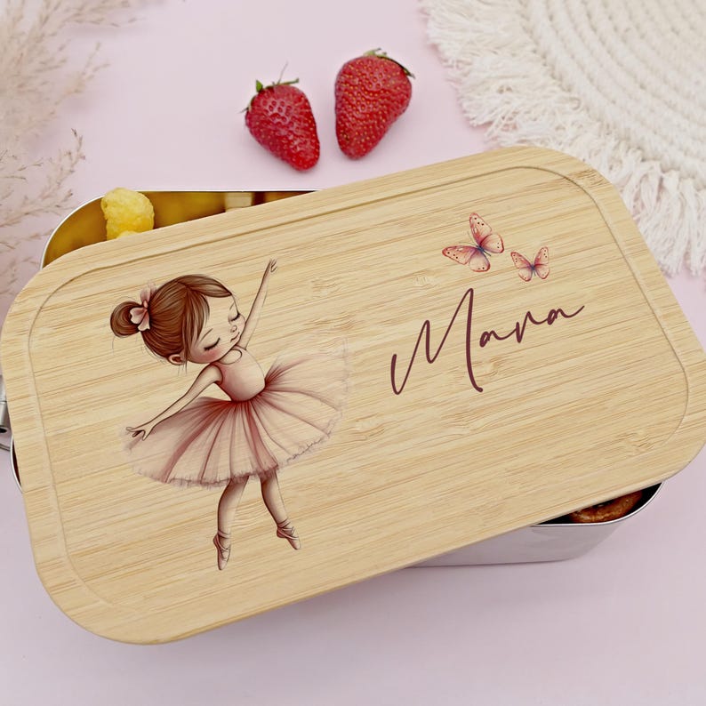 Pu&ograve; includere: Un portapranzo rettangolare con coperchio in legno chiaro. Il coperchio presenta un'illustrazione di ballerina con tut&ugrave; rosa, due farfalle e il nome "Mana" in corsivo. Il portapranzo &egrave; aperto, rivelando cibo all'interno.