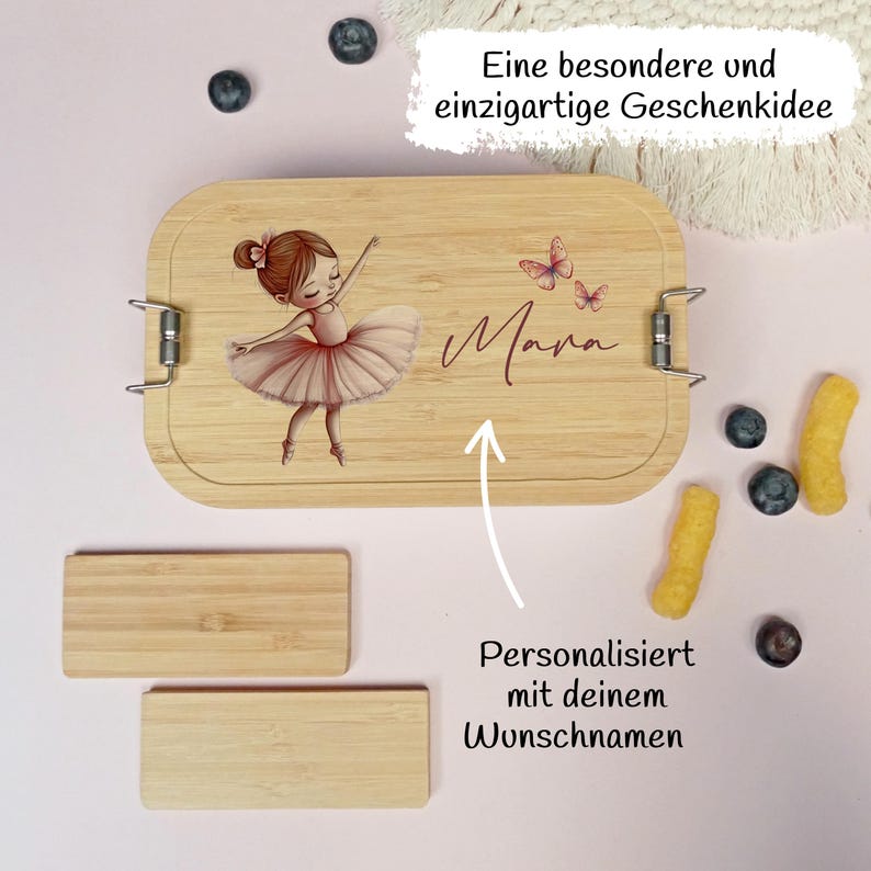 Pu&ograve; includere: Un portapranzo in legno chiaro con un'illustrazione di ballerina e il nome "Mara". La scatola ha chiusure in metallo e due piccoli pezzi rettangolari di legno. Il testo sulla scatola recita "Personalizzato con il tuo nome desiderato".