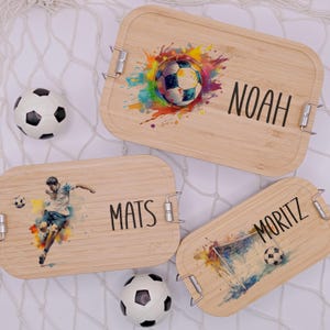 Puede incluir: Tres fiambreras de madera con diseños de acuarela coloridos que representan balones de fútbol, un jugador de fútbol y una portería. Las fiambreras tienen cierres metálicos y están personalizadas con los nombres Noah, Mats y Moritz.