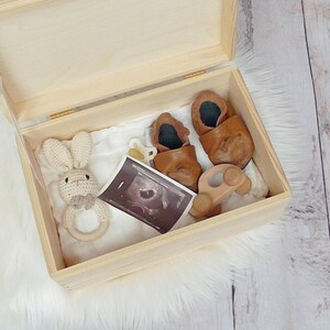 May include: A wooden keepsake box with a white fabric lining. The box contains a crocheted bunny rattle, a pair of brown baby shoes, a wooden toy, and a black and white ultrasound image.