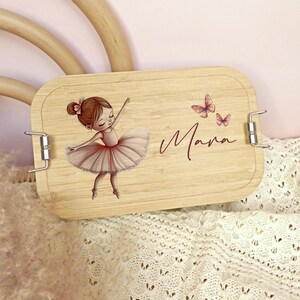 Pu&ograve; includere: Un portapranzo rettangolare in legno con un'illustrazione di ballerina e il nome "Mara" in corsivo. La ballerina indossa un tut&ugrave; rosa e ha le braccia tese. Sono raffigurate anche due farfalle rosa.