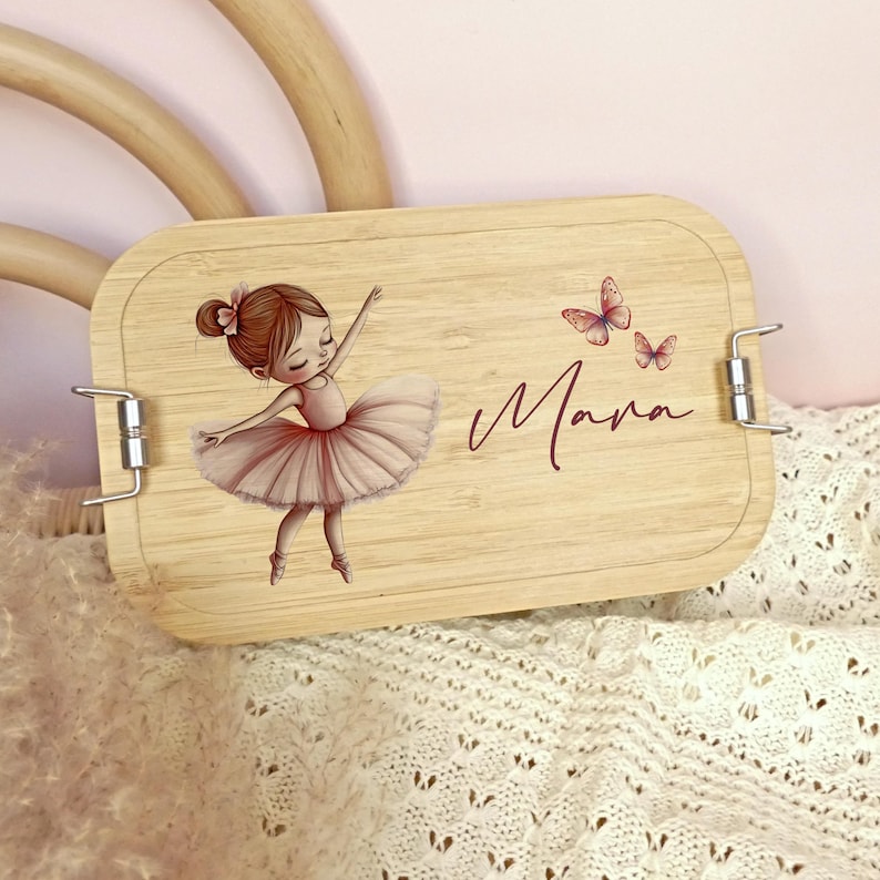 Pu&ograve; includere: Un portapranzo rettangolare in legno con un'illustrazione di ballerina e il nome "Mara" in corsivo. La ballerina indossa un tut&ugrave; rosa e ha le braccia tese. Sono raffigurate anche due farfalle rosa.