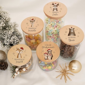 Könnte beinhalten: Fünf durchsichtige Gläser mit hellen Holzdeckeln, die jeweils mit einer Illustration eines Waldtieres mit Weihnachtsmütze und kleinen Sternen bedruckt sind. Die Gläser sind mit verschiedenen bunten Süßigkeiten und Schokolade gefüllt.