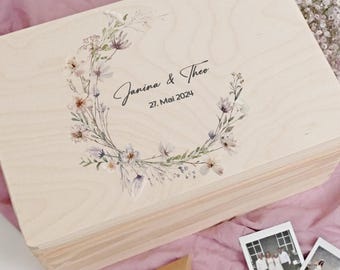 Op de afbeelding: Een lichtgekleurde houten kist met een bloemenkransontwerp en de namen "Janina & Theo" en de datum "27. Mai 2024" op het deksel. De kist is rechthoekig en waarschijnlijk een herinnerings- of souvenirkist.