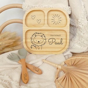 Può includere: Un piatto diviso in legno con un design di leone, sole e cuori. Il piatto è personalizzato con il nome "Paul" e il testo "Hier krümelt".
