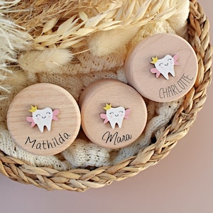 Op de afbeelding: Drie ronde houten dozen met een tandontwerp, elk met een naam: "Mathilda", "Mara" en "Charlotte". De tand heeft een gele kroon en roze vleugels. De dozen liggen in een gevlochten mand met gedroogde planten en een witte gebreide deken.