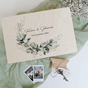 Può includere: Un cofanetto in legno con un design floreale e i nomi "Tamara & Johannes" e la data "02. Novembre 2024" stampati sul coperchio. L'immagine include foto polaroid, una chiave e una piccola scatola regalo.