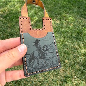May include: A brown and green leather card holder with a stitched border and a leather strap. The front features an engraved image of a person riding a horse in a landscape with mountains.