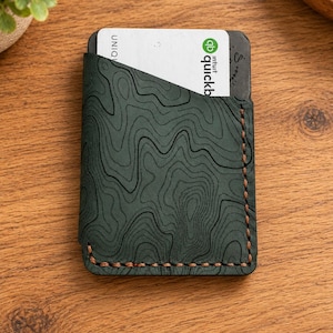 May include: A dark green leather card holder featuring a black topographic map pattern embossed on the surface. The wallet is constructed with visible brown stitching along the bottom and side edges and contains a white Intuit QuickBooks payment card.