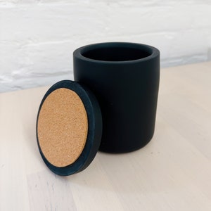 Könnte beinhalten: Eine schwarze Keramikdose mit einem Korkdeckel. Die Dose steht auf einer hellen Holzoberfläche.