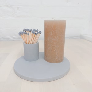 May include: A grey ceramic candle holder with a round base and a cylindrical top. A beige candle is placed on the holder. A small grey ceramic container with a bunch of matches is placed on the holder.