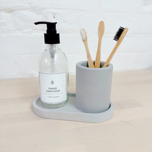May include: A grey ceramic bathroom organiser with three bamboo toothbrushes and a clear glass bottle with a black pump dispenser labelled "HAND SANITIZER I am reusable".