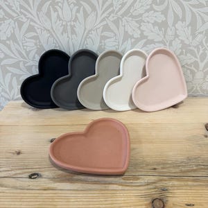 Può includere: Una collezione di vassoi a forma di cuore in vari colori, tra cui nero, grigio, beige, bianco e rosa. Un vassoio color terracotta in primo piano. I vassoi sono disposti su una superficie di legno, con una carta da parati floreale sullo sfondo.